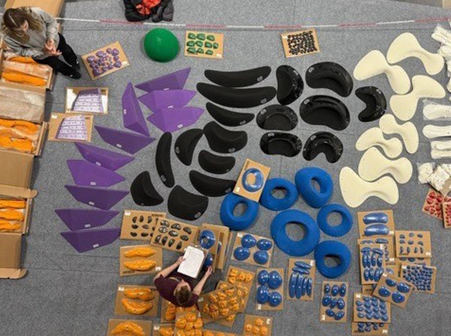 Overhead view of a layout of indoor rock climbing holds spread across a carpeted floor, including white wavy ergonomic holds, black curved holds, purple triangular volumes, blue circular and ring-shaped slopers, with two people sorting them. 