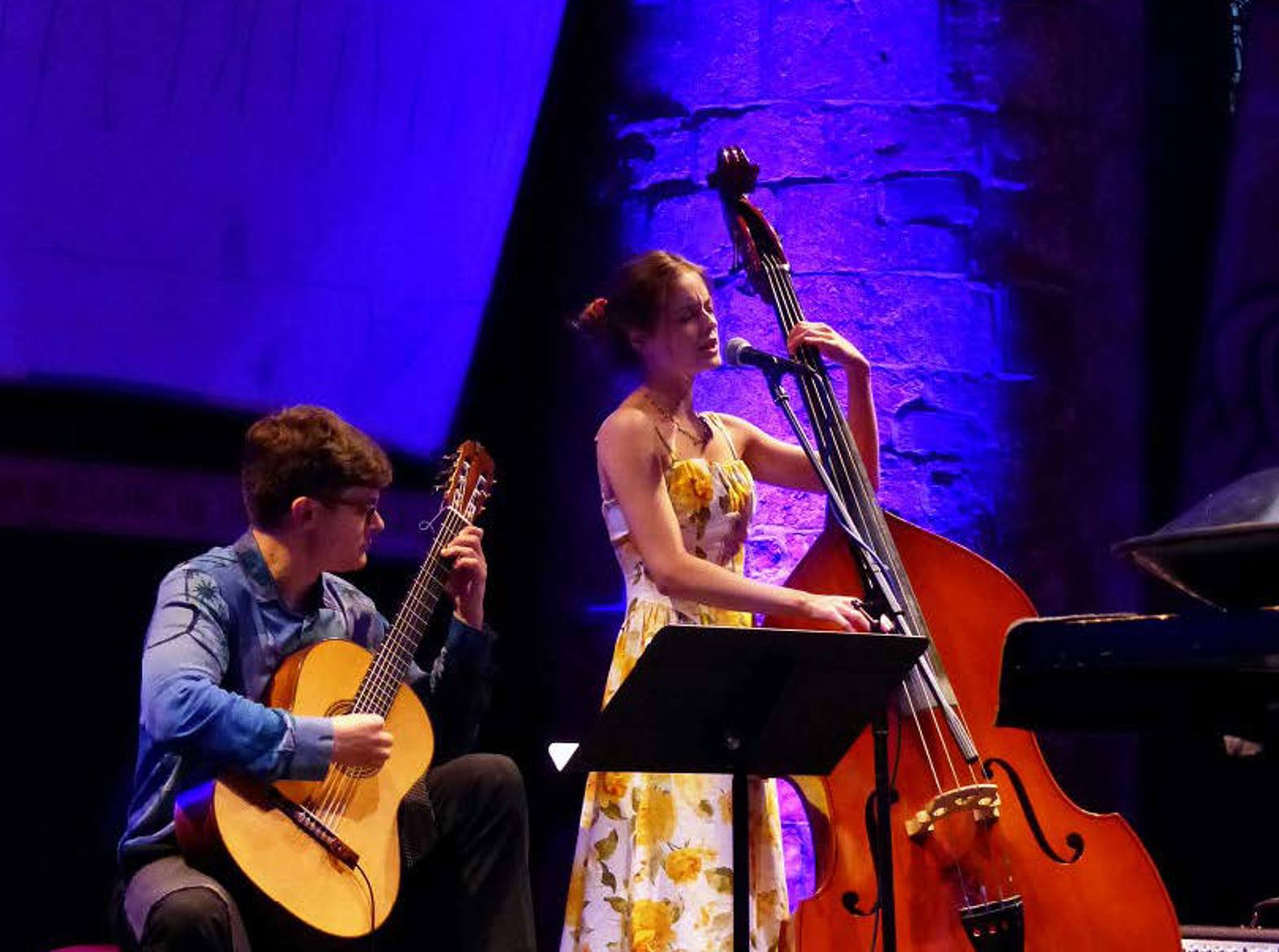 Two musicians perform on stage under blue and purple lighting: a guitarist holding an acoustic guitar and a double-bass player  stands beside. 