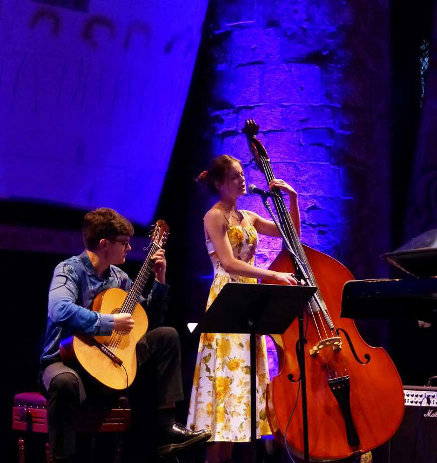 Two musicians perform on stage under blue and purple lighting: a guitarist holding an acoustic guitar and a double-bass player  stands beside. 