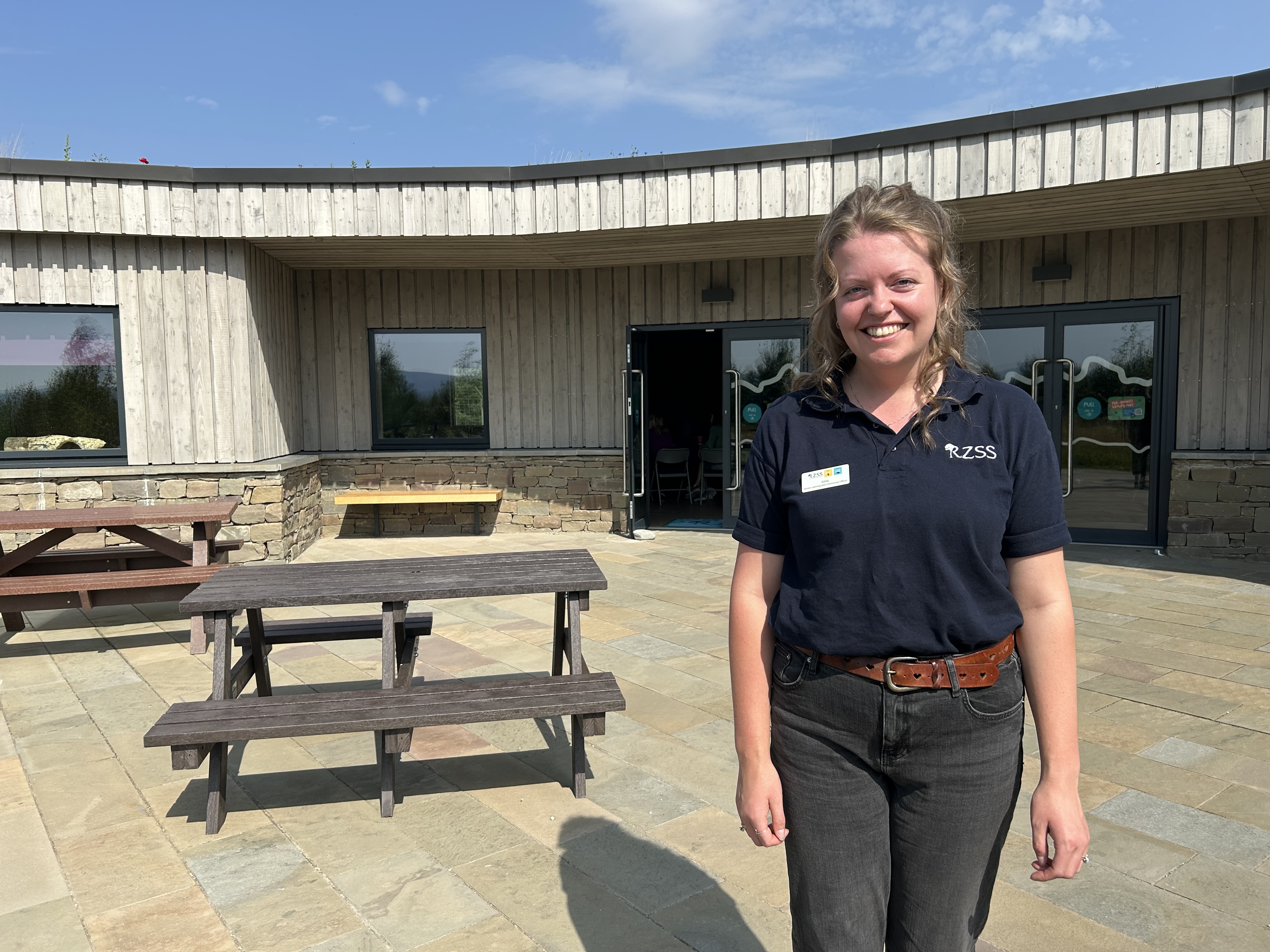 A woman stands and smiles in front of a modern wooden building with large windows. She is wearing a navy blue polo shirt with the RZSS logo and dark jeans. The area is paved with stone tiles and has picnic tables. The weather is sunny with a blue sky and light cloud.