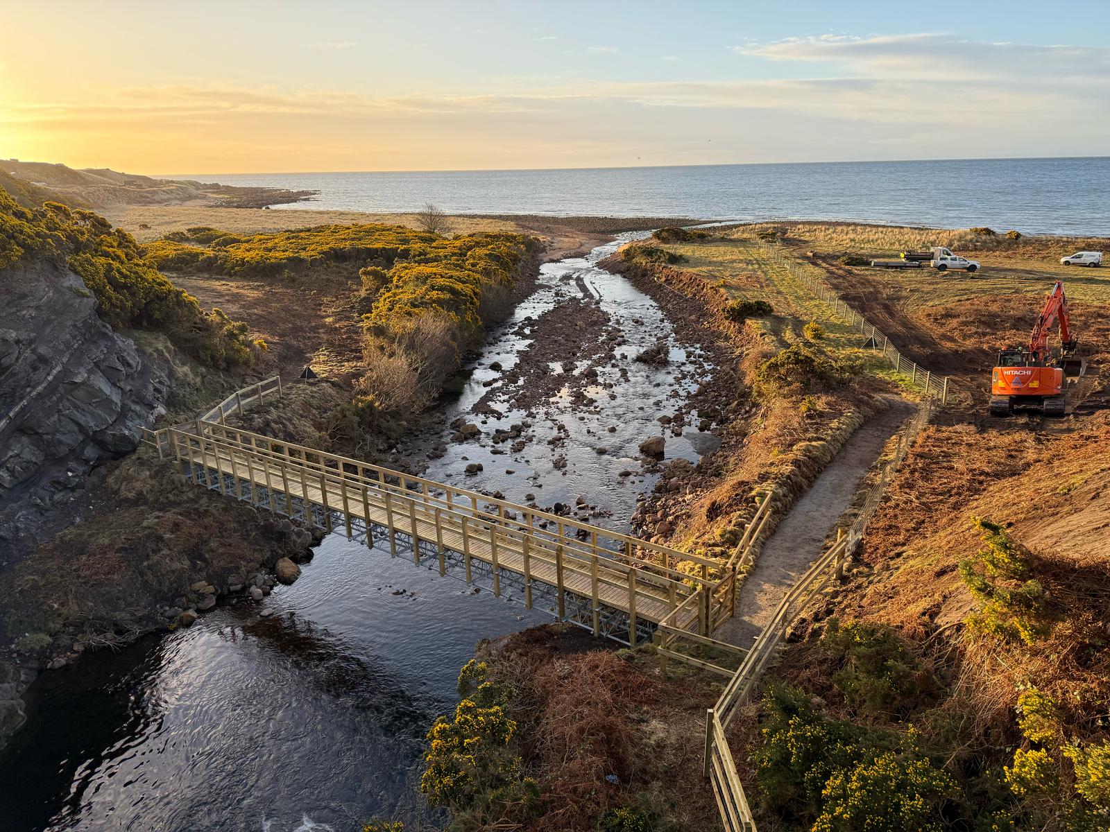 In the foreground, a wooden bridge spans a burn. The background shows the sun setting on a stretch of coast. 