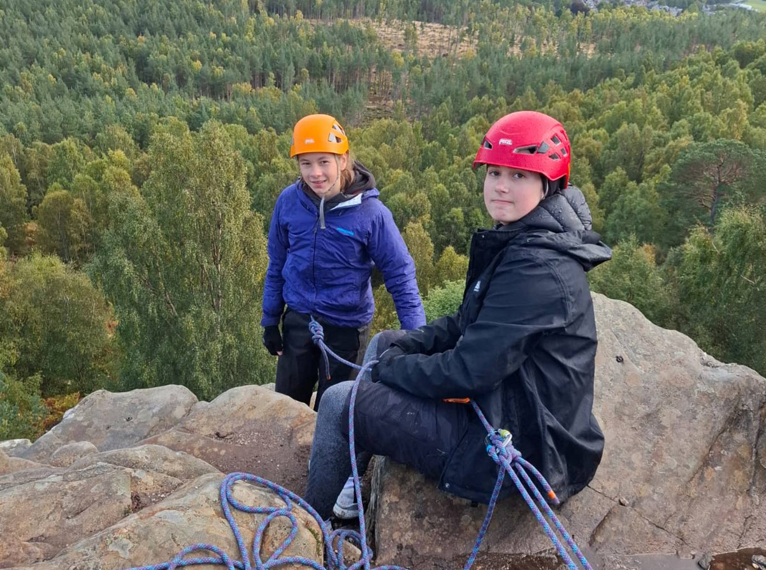 Two climbers wearing helmets and outdoor jackets sitting on a rocky cliff edge with climbing ropes coiled in front of them. Behind them stretches a vast landscape of forest, fields, and distant hills beneath a cloudy sky.