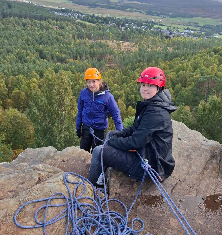 Two climbers wearing helmets and outdoor jackets sitting on a rocky cliff edge with climbing ropes coiled in front of them. Behind them stretches a vast landscape of forest, fields, and distant hills beneath a cloudy sky.