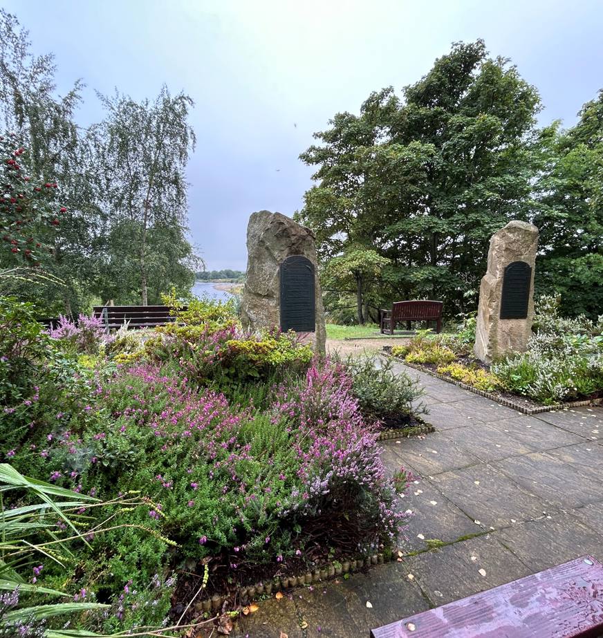 A picnic bench, surrounding by purple heathers and other bedding planrs, sits amid a riverside garden. 