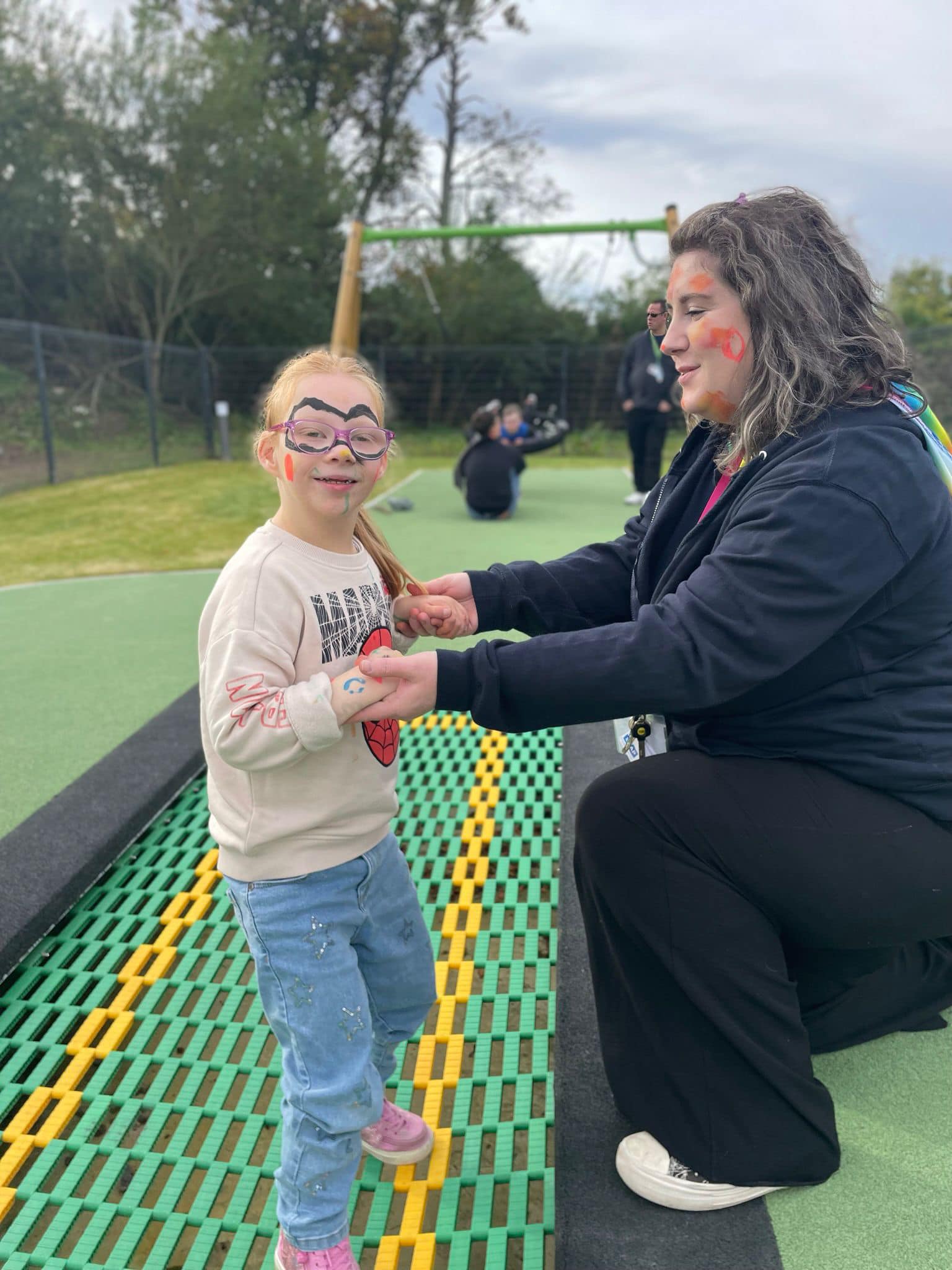 A young girl, who has her face painted like a super hero, is standing on a trampoline. A woman kneels beside her, holding her hands. 