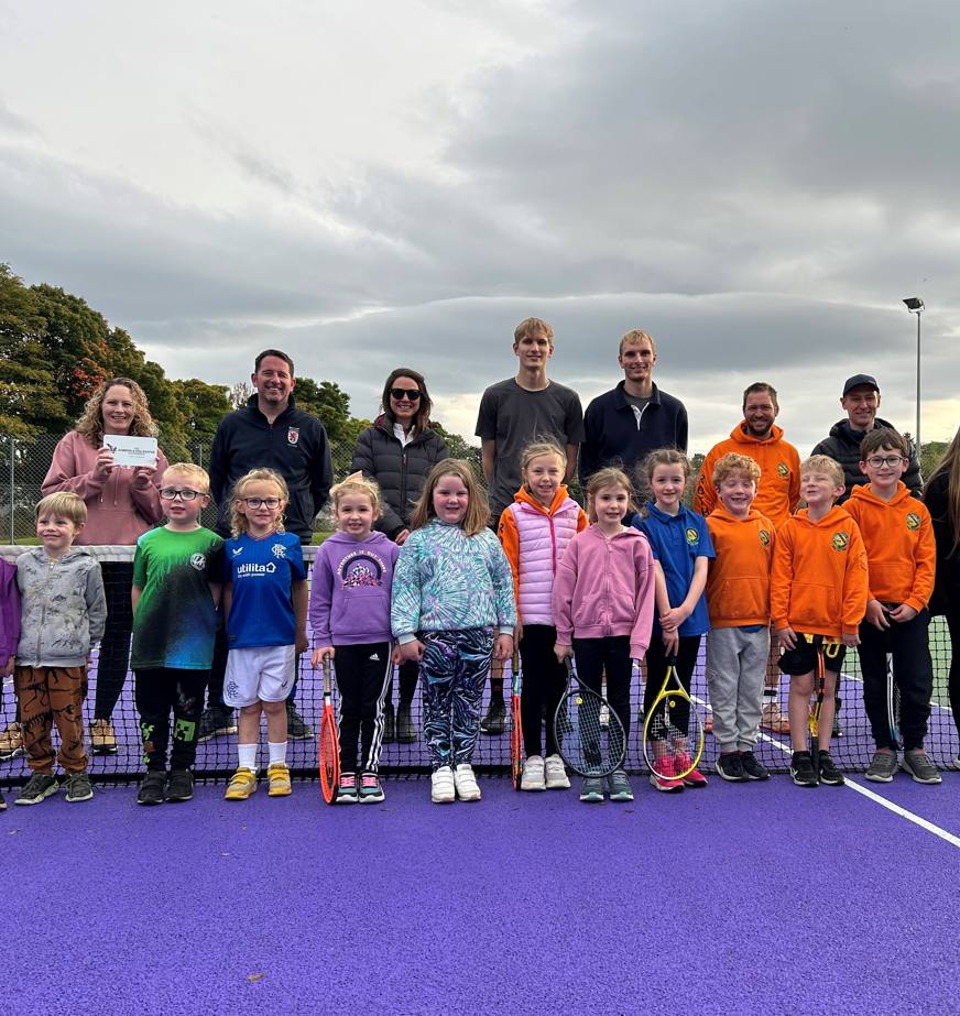A group of children and adults stand in a group in the centre of a tennis court, next to the net. The courts surface is purple and the surroundings are green. 